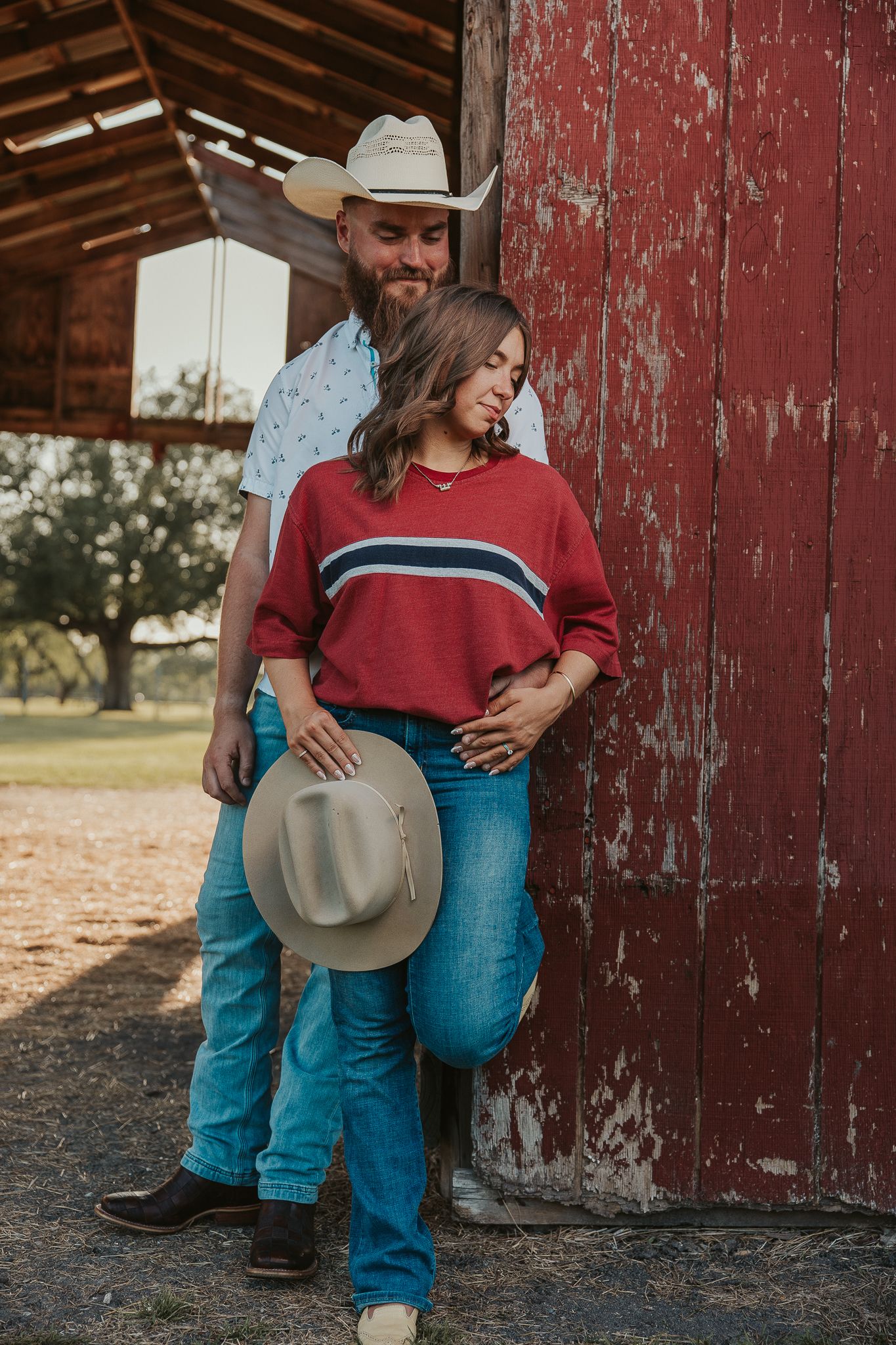 Couple in cowboy hats leaning against a rustic red barn door, wearing jeans and western attire on a farm.