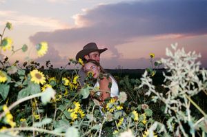 Couple embracing romantically in sunflower field at purple sunset, man wearing cowboy hat.