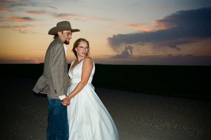 Bride and groom hold hands at sunset, groom in cowboy hat and jeans, bride in flowing white gown.