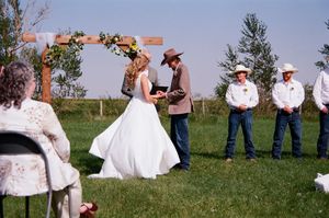 Outdoor rustic wedding ceremony under wooden arch with cowboy-themed groomsmen on sunny day.