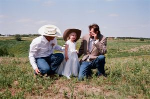 Two men crouching beside young girl in white dress in sunny green meadow, one wearing cowboy hat.