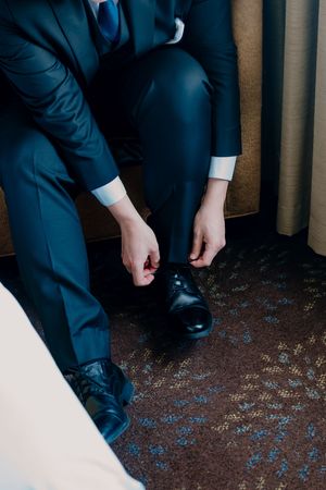 Man in navy blue suit tying black dress shoes while seated, preparing for formal event.