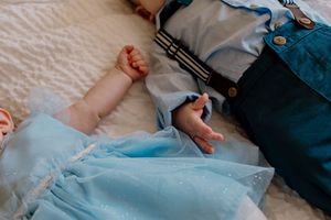 Two babies lying down, one in a blue dress and one in denim overalls, holding hands tenderly.