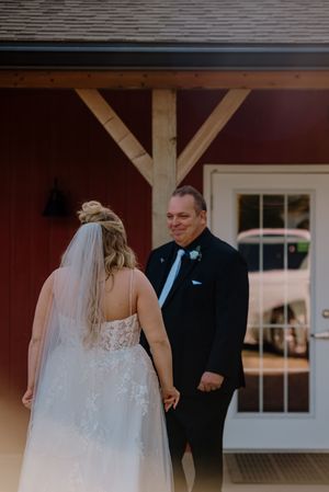 Father smiles warmly at bride in lace wedding dress and veil during first look outside a rustic red barn venue.