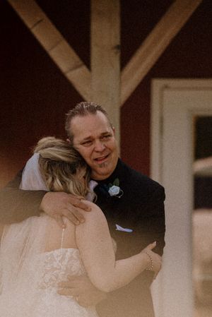 Emotional groom hugging bride in wedding attire outside rustic red barn venue.