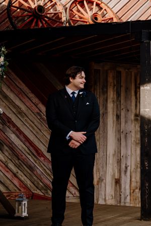 Groom in black suit with blue tie smiling at barn wedding venue with rustic wooden walls and wagon wheel decor.