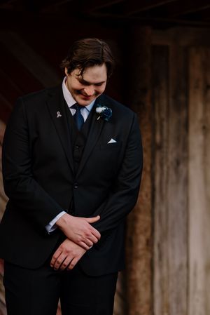 Groom in black suit with blue boutonniere looking down emotionally during wedding ceremony at rustic barn venue.