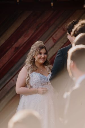 Smiling bride in lace wedding gown during outdoor ceremony with rustic red barn backdrop.