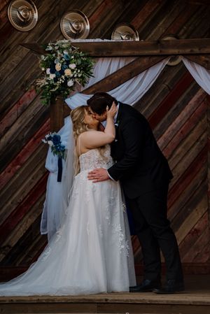 Bride and groom share first kiss at rustic barn wedding altar adorned with blue and white floral arrangements.