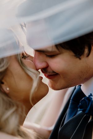 Romantic close-up of bride and groom almost kissing under wedding veil, groom wearing navy bow tie.