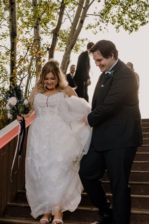 Bride in lace gown descends wooden stairs with groom in black tuxedo at outdoor wedding ceremony.