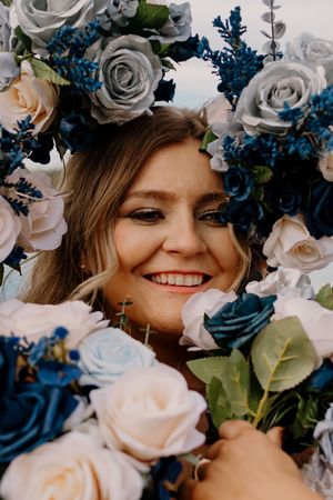 Smiling bride surrounded by blue, white, and gray roses, wearing a floral crown at her wedding ceremony.