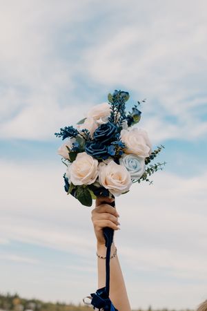 Hand holding a bridal bouquet of white and blue roses against a cloudy sky.