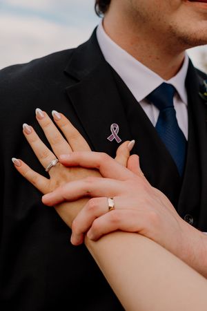 Close-up of newlyweds hands showing wedding rings with pink ribbon pin on grooms black suit.