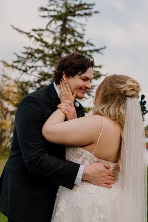 Groom in black tuxedo embraces bride in lace wedding gown outdoors, sharing an intimate moment near evergreen trees.