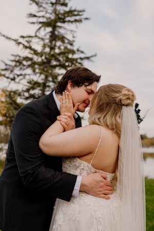 Groom tenderly kisses bride in lace wedding gown outdoors near evergreen trees.