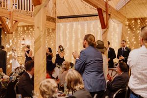 Wedding guests applaud during reception speeches in elegant barn venue with fairy light backdrop.