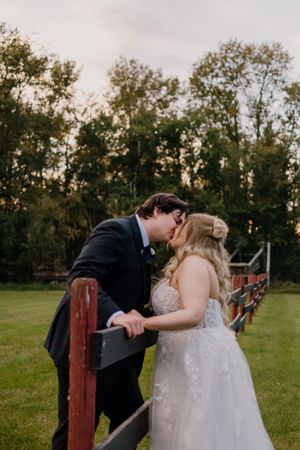 Bride and groom share romantic kiss leaning on rustic red fence at outdoor wedding venue.