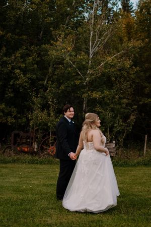 Bride in lace gown and groom in black suit hold hands walking through lush green garden.