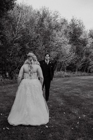 Bride in lace ball gown walks toward smiling groom in black suit on a grassy outdoor wedding venue.