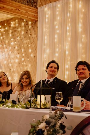 Wedding party smiles at head table adorned with greenery and candles against a fairy light backdrop.