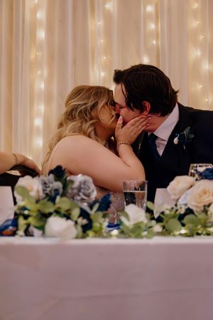 Bride and groom share a romantic kiss at their wedding reception sweetheart table adorned with white roses.