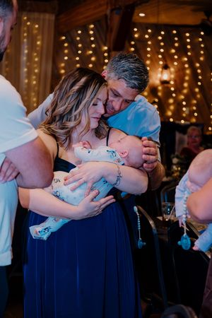 Couple in navy attire shares a tender moment with a newborn baby at a warmly lit barn reception.