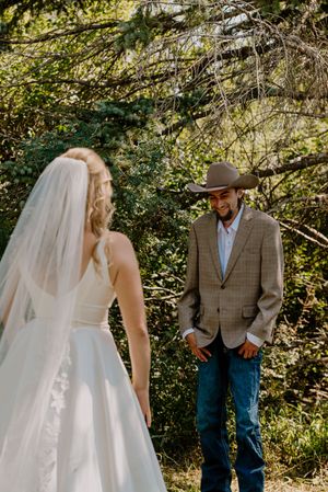 Groom in cowboy hat and plaid blazer smiling at bride in white wedding dress during outdoor first look.