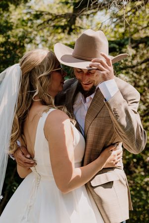Emotional groom in cowboy hat and tan blazer wiping tears as bride in white dress and veil embraces him outdoors.