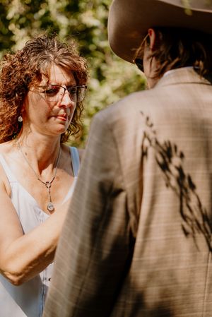 A bride with curly hair and glasses exchanges vows with a groom in a tan suit and cowboy hat outdoors.