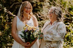 Bride in white gown holding colorful bouquet smiles with older woman in floral jacket outdoors at wedding ceremony.