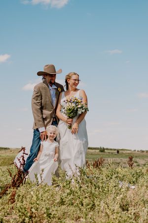 Couple in wedding attire with young girl holding wildflower bouquet in open field.