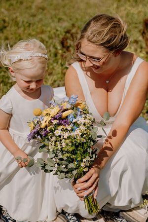 Smiling bride in white dress crouches to share colorful wildflower bouquet with young flower girl outdoors.