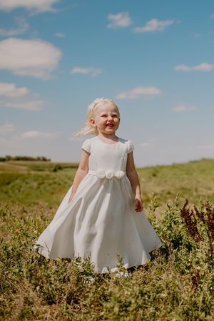 Blonde toddler girl in white flower girl dress standing in green field under blue sky.
