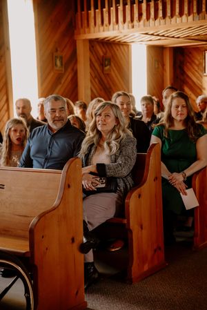 Wedding guests seated in wooden church pews during a ceremony, with a woman in a gray blazer prominently featured.
