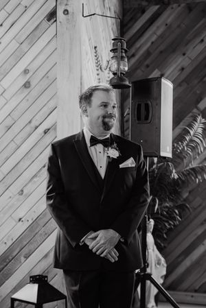 Groom in black tuxedo with bow tie and boutonniere smiling at wedding ceremony in rustic barn venue.