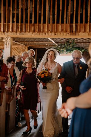 Bride walks down the aisle in a barn wedding venue, escorted by parents, holding a vibrant bouquet of orange and yellow flowers.