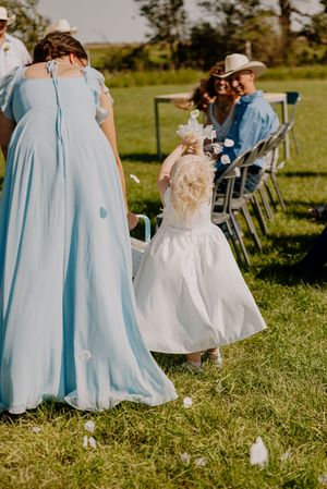 Flower girl in white dress walks down outdoor wedding aisle beside bridesmaid in light blue gown.