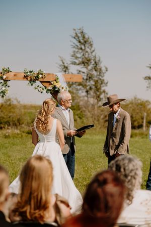 An outdoor wedding ceremony with a bride, officiant, and groom in a cowboy hat beneath a floral arch.