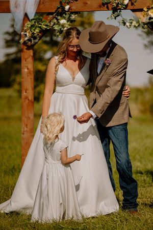 Bride and groom in cowboy hat receive flower from flower girl at outdoor rustic wedding ceremony.