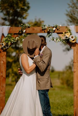 Bride and groom share first kiss under rustic wooden floral arch at outdoor wedding ceremony.