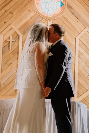 Bride and groom share their first kiss at the altar in a rustic wooden chapel with a stained glass window and cross.
