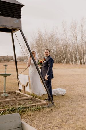 Bride and groom posing outdoors near a rustic swing structure in a field with bare birch trees during a fall wedding.
