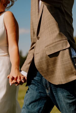 Bride and groom hold hands in matching white dress and brown plaid blazer with jeans outdoors.