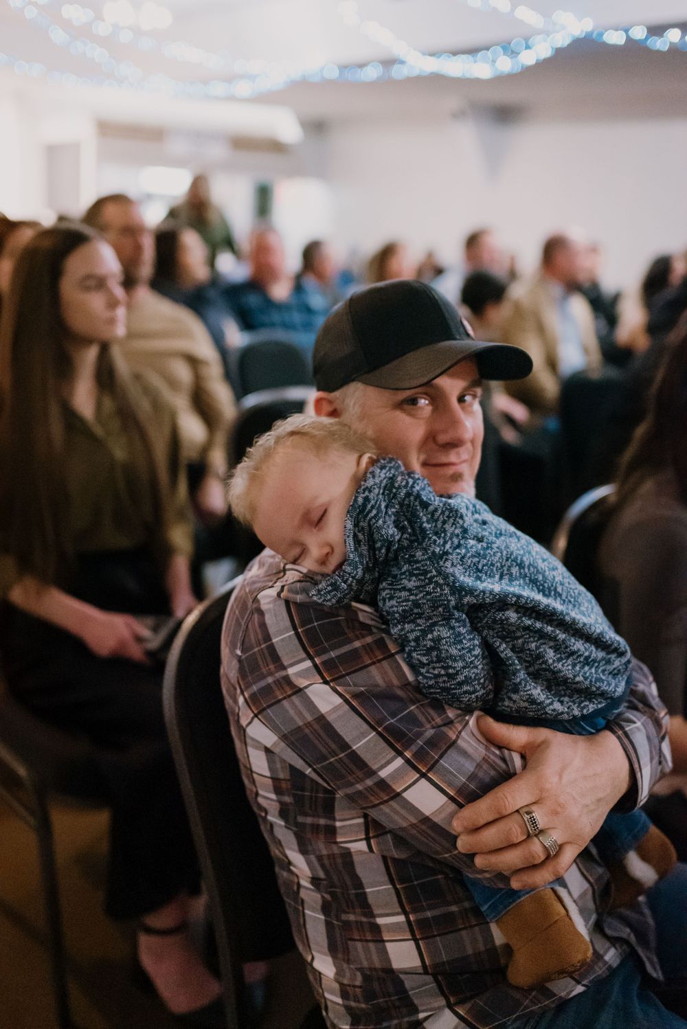 Man in plaid shirt and black cap holds sleeping toddler at an indoor gathering, smiling warmly at camera.