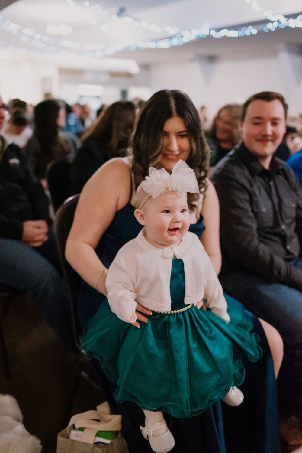 A smiling baby in a green tutu dress with a white bow headband held by a woman at a wedding reception.