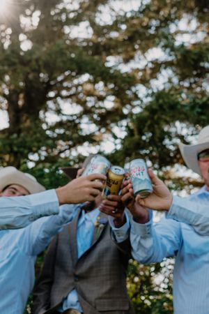 Groomsmen in cowboy hats toasting with Coors Light beer cans outdoors at a country wedding celebration.