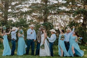 Wedding party in cowboy hats, bridesmaids in blue dresses with jeans, celebrating outdoors among trees.