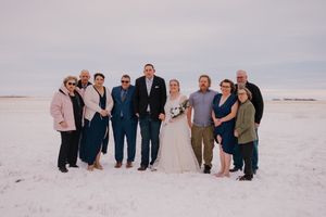 Wedding party posing outdoors in snowy winter landscape, bride in white gown with bouquet surrounded by family in formal attire.