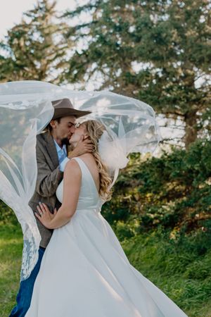 Bride and groom kiss under a flowing veil billowing in the wind outdoors.
